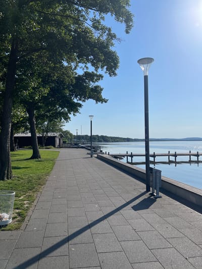 This image depicts a paved walkway along the shore of a lake in Wunstorf, Germany, on a clear, sunny day. The walkway is constructed of large, grey square tiles and is bordered on the left by a grassy area with trees and a small wooden building. On the right, the walkway overlooks the calm lake, with a wooden pier extending into the water. A modern lamppost stands on the right side of the walkway, casting a long shadow across the tiles. In the foreground on the left, a wire trash receptacle is visible. In the background, the treeline of the opposite shore is visible, along with a clear blue sky. The scene appears serene and unoccupied, with no people or discernible activity.