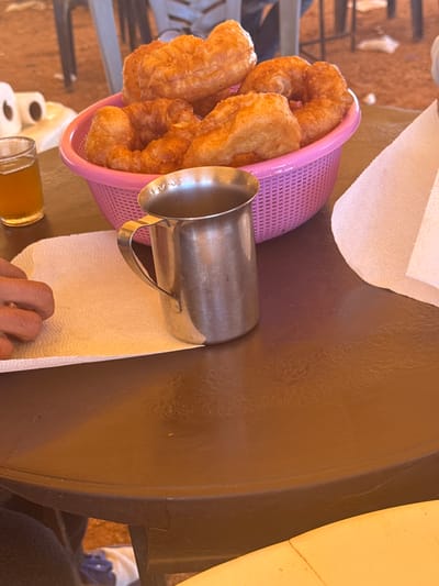 A high-angle shot of a table in Boufakrane, Morocco. A pink plastic basket filled with fried pastries rests on the edge of the table next to a stainless steel pitcher. A roll of paper towels sits on the table, with a person's hand reaching for a sheet. A small glass container with a golden liquid sits near the hand. The table is brown and appears to be plastic. The environment suggests a casual, possibly outdoor dining setting. A person wearing a blue medical glove is seen in the background.