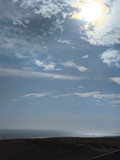 The image shows a daytime view of the ocean near Huanchaco, Peru. The sky is mostly blue with scattered clouds, and the sun shines brightly from the upper right, partially obscured by clouds. Below the sky, a wide expanse of the ocean stretches toward the horizon, reflecting sunlight on its surface. The foreground features a dark, grassy area possibly a cliff or a hill overlooking the sea. A fence or railing is visible along the edge of the land, separating it from the water. The lower right corner of the image reveals a portion of what appears to be a road.