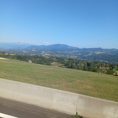 This is a high-angle, wide shot capturing a scenic overlook of the landscape surrounding Gap, France. In the foreground, a concrete barrier runs along the bottom of the frame, separating the viewpoint from a paved road. Beyond the barrier, a gently sloping grassy field occupies the midground. Further into the distance, a valley unfolds with rolling hills covered in a patchwork of forests and agricultural fields. In the far background, a range of mountains, some with snow-capped peaks, forms a majestic backdrop under a clear blue sky. A small, light-colored building with a dark roof is nestled amongst the trees in the mid-distance to the left of center. The overall scene depicts a serene, rural vista bathed in bright sunlight.