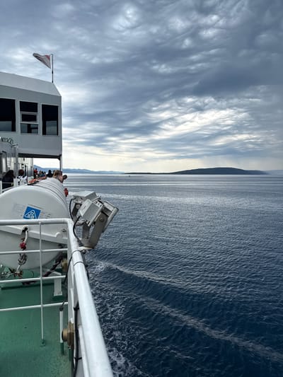 The image captures a view from a ferry, likely departing from Drvenik, Croatia. The foreground features the side of the ferry, including a white railing, a green deck, and a large white life raft with visible labels. Mounted on the railing are two grey floodlights with black cables. In the background, a calm, deep blue sea stretches towards a distant, low-lying island or coastline under a cloudy, overcast sky. The clouds are textured with grey and white, suggesting a dramatic or moody atmosphere. Several people are visible on the deck of the ferry, leaning over the railing and looking out at the sea. A flag with a red and white design flies from a pole on the upper structure of the ferry. The overall scene conveys a sense of travel and the vastness of the ocean.