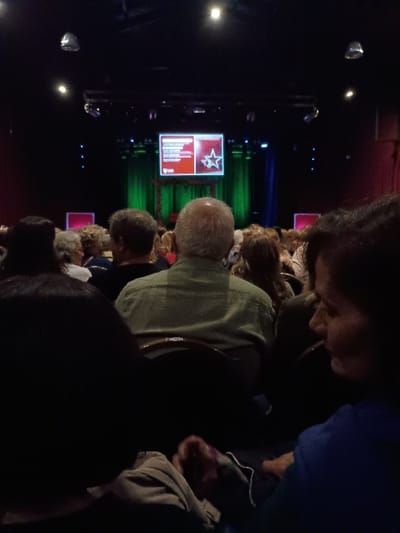The image shows a dimly lit auditorium with a large screen displaying a presentation. The screen shows a red and white logo. The audience is seated in rows, facing the screen. The main subject of the image is a man in a green shirt. He sits in the front row. There are other people in the audience, but they are not identifiable. The environment is dark, except for the screen and the overhead lights. The location of the scene is an auditorium in General Pacheco, Argentina, at the coordinates (-34.457, -58.633). The people in the audience are observing the presentation on the screen. There is no visible interaction among the people in the audience.