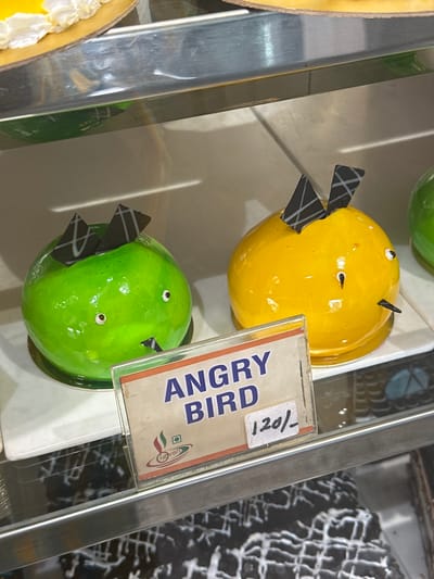 In Mathura, India, a display case contains pastries, including two shaped like Angry Birds, one yellow and one green, along with a price tag of 120/-.