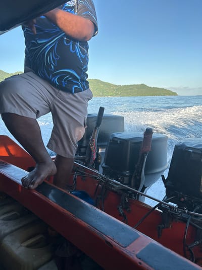 This is a close-up, low-angle shot of a person standing on a red and black boat, with two outboard motors visible in the background. The person, whose lower body is in focus, is wearing light-colored shorts and a black t-shirt with a blue and white floral pattern. Their right foot is bare and placed on the edge of the boat. The boat is in motion, indicated by the white spray and wake from the water. In the distance, a lush green island with hills slopes down to the ocean. The sky is clear and blue. The scene is likely at sea near the islands of Tidore, Indonesia, given the tropical island landscape.