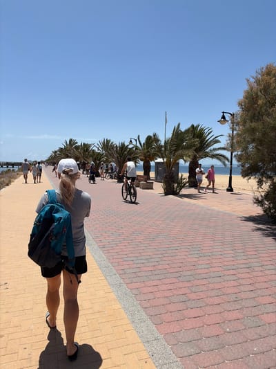 The image captures a sunny day along the waterfront promenade in San Pedro del Pinatar, Spain. In the foreground, a woman with blonde hair tied back, wearing a white cap, grey t-shirt, and black shorts, walks away from the camera, carrying a teal backpack. She is on a yellow brick-paved walkway. To her right, the promenade transitions to red and gray brick paving.

Further along the promenade, several people are visible. A man on a bicycle rides past a row of palm trees. Other individuals are seen walking or standing near the beach, which is to the right of the promenade and separated by a sandy area and more palm trees. A black lamppost stands on the right side of the image, with a tree to its right. The sky is clear and blue, and the sea is visible in the background. A pier or jetty extends into the water on the far left. The overall scene depicts a leisurely day out in a coastal town.