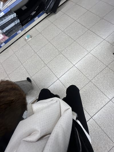 The scene captures a downward angled view of a tiled floor, meeting a shelf displaying folded clothes. The focus shifts to a person’s lower half, clad in black leggings and a black jacket trimmed with white. Overlapping these are folds of a dotted beige fabric, and a brown fur item partially obscuring the lower view. The composition has a casual, observational feel, likely taken from a shopper's perspective in a retail environment.