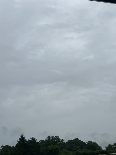 The image shows a view of a sky filled with overcast, grey clouds. The lower portion of the image features a dense line of dark green trees, with the top of some evergreen trees being visible above the deciduous trees. The scene appears to be an outdoor environment, likely a suburban or rural area, given the presence of the trees. There are no people or any visible structures apart from what could be a dark, horizontal line along the very top edge of the image, which might indicate the edge of a building or a roofline. The overall atmosphere is muted due to the cloudy sky. The location is described as Astomi, Latvia.