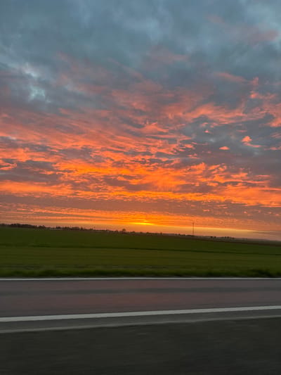 The image shows a landscape scene at dusk, likely taken on a road in or near Annœullin, France. A vibrant sunset dominates the sky, with fiery orange and pink clouds streaking across the horizon. To the right of the frame, a green field stretches into the distance, and a gray asphalt road with white lane markings occupies the foreground. There are no visible people or interactions in the scene. A distant utility pole can be seen on the horizon. The image is oriented sideways.