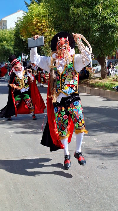 In the city of Wanchaq, Peru, on a sunlit day, two performers in elaborate traditional costumes are captured mid-stride on a paved street. The primary subject, in the foreground, is a dancer in a red and black outfit adorned with vibrant embroidered birds and flowers. They wear a striking mask with a red face, sharp teeth, and horns, topped with a black hat. The dancer holds a gray rectangular object in their left hand and a braided rope whip in their right, appearing to be in mid-jump, with their feet in black and red shoes and white leggings airborne. Behind them, another masked performer in a similar costume, featuring a red cape and an embroidered vest, walks on the street. The background shows a tree-lined sidewalk with spectators sitting and standing, and a white building. The overall scene depicts a lively cultural procession or festival.
