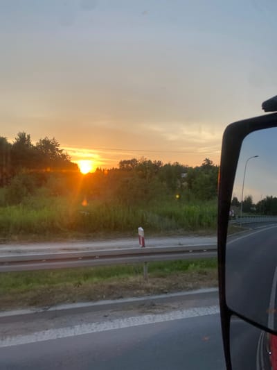 The image captures a sunset viewed from a vehicle, likely a truck, as indicated by the large side mirror in the foreground reflecting the sky and surrounding landscape. The sun, a bright orange orb, is setting behind a line of trees and lush green vegetation, casting lens flares and a warm glow across the scene. A road is visible in the lower right portion of the image, curving away from the viewer and bordered by a metal guardrail and a grassy verge. A white and red-striped marker post stands near the guardrail. The sky transitions from a pale blue at the top to a gradient of orange and yellow near the horizon. The scene appears to be set in a rural or semi-rural area outside of Zakrzów, Poland, given the reflective elements of the mirror and the roadside markers.
