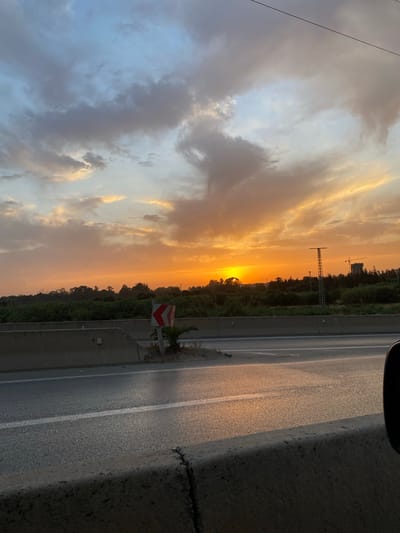 The image shows a scenic view of a highway at sunset in El Karia, Algeria. The sky is filled with dramatic clouds tinged with orange and yellow hues from the setting sun, which is partially obscured by trees on the horizon. A concrete barrier runs along the side of the highway, separating it from a strip of green vegetation. A traffic sign with red and white chevrons indicating a curve is visible on the left side of the road. In the background, there are more trees and a few structures, including what appears to be a tall tower or mast and a building with a crane nearby, suggesting industrial or construction activity. The asphalt road is wet and reflects the warm colors of the sky. The overall scene captures a tranquil moment of a sunset over a highway environment.