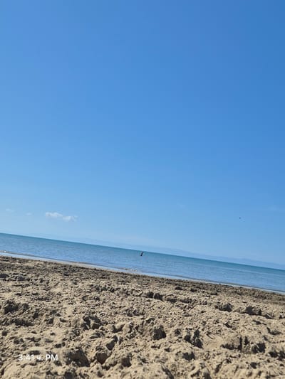 The image shows a close-up view of a sandy beach with the calm blue sea in the background under a clear blue sky. The sand is textured with ripples and small mounds. In the distance, a faint landmass is visible along the horizon. There is a single small red object in the water, possibly a buoy or a person wearing a red shirt. A very small dark object, possibly a bird or an insect, is visible high in the sky. The time stamp "3:41 ч. PM" is present in the lower left corner of the image. The scene depicts a tranquil beach on a sunny day, likely located in the city of Barletta, Italy, given the context.