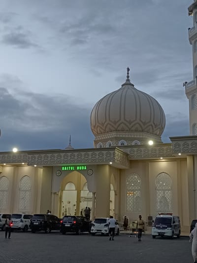In the city of Mee Tanjong Usi, Indonesia, a mosque named "Baitul Huda" is depicted at dusk. The mosque's prominent dome, adorned with intricate patterns and topped with a crescent moon and star finial, dominates the upper half of the frame. A minaret is visible to the right of the dome. The mosque's facade features a wide entrance, ornate architectural details, and patterned windows. In front of the mosque, a paved area serves as a parking lot for several vehicles, including cars and a white ambulance. A few individuals are present, some walking and others near the vehicles. The sky is overcast, with dramatic clouds adding to the atmospheric quality of the scene.
