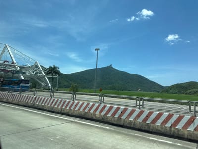 The image depicts a scenic view from a moving vehicle, likely a bus, in Hoa Xuan, Vietnam. The foreground shows a concrete road with white lane markings. A concrete barrier with alternating red and white diagonal stripes separates the road from a walkway or median. Beyond the barrier, a metal fence with a diamond pattern is visible, and further back, a bright green rice field stretches towards lush, tree-covered mountains.
In the background, a prominent, pointed mountain peak stands against a clear blue sky dotted with a few wispy white clouds. A tall lamppost with multiple lights is situated in front of the mountain. To the left, a white, modern-looking structure, possibly part of a bridge or overpass, is visible with a red life preserver hanging from it. A blue bus is partially visible under this structure. A single palm tree stands near the structure.
The time of day appears to be midday, given the bright sunlight and clear sky. The weather seems to be warm and sunny, ideal for outdoor activities. The presence of the rice field, mountains, and tropical vegetation suggests a rural or semi-rural setting. There is no discernible activity taking place other than the travel from the perspective of the camera.
Visible text includes "8 m" on a red circular sign on the left. Additionally, a yellow triangular sign with a black exclamation mark and some Vietnamese text is visible near the lamppost, indicating a warning. The overall mood conveyed is one of peaceful travel through a beautiful landscape.