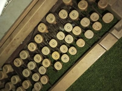 A close-up shot displays the keyboard of a vintage typewriter. The round, off-white keys feature faded letters and numbers. The arrangement appears in a standard QWERTY layout, with additional keys for numerals and symbols. A few keys are labeled with letters 'E', 'R', 'T', 'Y', 'U', 'I', 'O', 'P', 'A', 'S', 'D', 'F', 'G', 'H', 'J', 'K', 'L', 'Z', 'X', 'C', 'V', 'B', 'N', 'M'. The typewriter's frame is light brown, and green felt is visible between the keys.