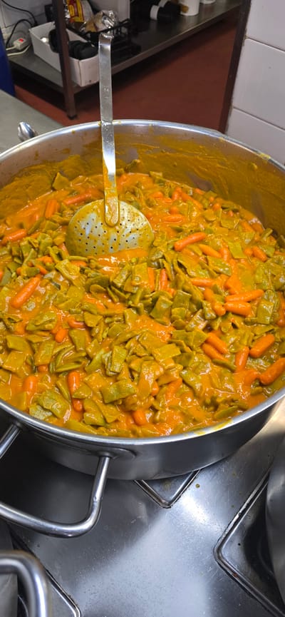 This media file captures a close-up, overhead view of a large pot filled with a vibrant, orange-hued stew containing green, flat beans and small, whole carrots. A metal ladle with numerous perforations rests partially submerged in the stew, suggesting it is being served or prepared. The pot itself is made of stainless steel and appears to be in a professional kitchen environment, indicated by the surrounding stainless steel surfaces and tiled walls.

The lighting suggests it could be daytime, given the brightness of the scene, though no direct sunlight or specific time of day cues are evident. The setting is likely a commercial kitchen or catering establishment.

Visible text is limited to a small "14" stamped on the metal handle of the ladle. There are no people visible in the frame. The overall impression is one of a hearty, freshly prepared dish ready to be served. The context provided, "Sallent de Gállego, Spain," places this scene in a specific geographic location, hinting at local cuisine.
