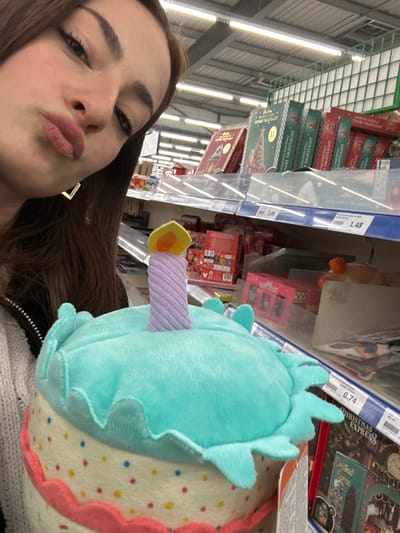 A young woman takes a selfie in a store aisle.  She's making a kissy face, and a plush toy shaped like a birthday cake with a turquoise frosting and a candle is positioned prominently. Shelves of various products are visible in the background, including "Diamond Painting NERY NIGHTLIGHT" boxes, "Christmas Express" items, and price tags displaying "2.99" and "1.48".