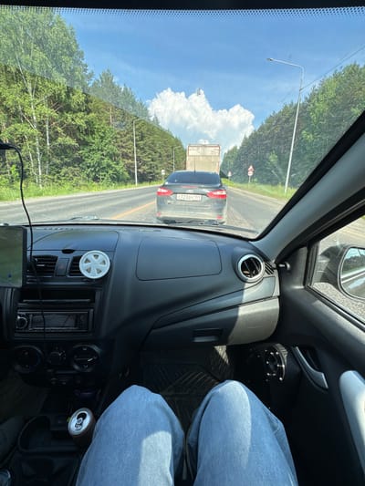 The image depicts a view from the driver's seat of a car traveling on a road lined with a dense forest of birch and pine trees under a bright, cloudy sky. The road appears to be a two-lane highway. Ahead, a dark gray Ford sedan is visible, its license plate reading "C723BY 159". Further ahead, a white truck with a beige cargo area is moving in the same direction. Several road signs are present along the roadside, including what appear to be speed limit or cautionary signs.
Inside the car, the dashboard is visible, featuring an air conditioning vent with a white decorative element, a car stereo, and a navigation device mounted on the left. The driver's legs, clad in blue jeans, are in the foreground. A can of soda is placed in a cup holder. The overall environment suggests a daytime journey through a rural or semi-rural area, possibly on a highway leading to or from Pesterevo, Russia, given the context.