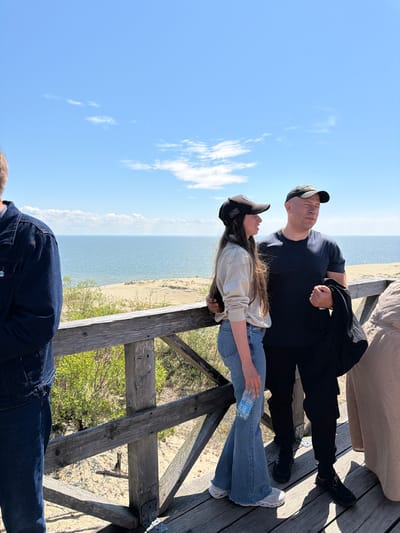 In Morskoye, Russia, on a wooden boardwalk overlooking a sandy dune landscape and the sea, a man and a young woman are standing. The man, wearing a dark t-shirt and a baseball cap, holds a black jacket in his right hand. The young woman, wearing a light-colored sweatshirt, jeans, and a baseball cap, is holding a bottle of water. To the left, the back of another person wearing a denim jacket is visible. Another person, mostly obscured, is visible on the far right. The sky is blue with scattered clouds, and the sea is calm.