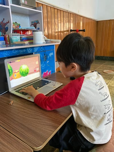 An indoor scene in the city of Đak Đoa, Vietnam, depicts a young child seated at a wooden desk, facing right, engaged with a silver MacBook laptop. The child has dark hair and wears a white raglan-style shirt with red sleeves and black outline drawings of dinosaurs labeled "TRICEROP," "THUNDER CR," "FLYING DIN," and "CYRATOS" on the back, along with dark pants. Their left hand rests on the laptop keyboard, with the index finger poised over a key. The laptop screen displays a game or application featuring two watermelons and one yellow fruit against a wooden textured background. To the left of the laptop, on the desk, a small golden-brown toy car is visible.
Behind the desk and to the left, a light blue and white shelving unit is present. The lower section of the unit features blue panels with white decorative drawings. The shelves hold various items, including a dark box on top, a green box and a blue container on the middle left, and a red box labeled "TOTAL," a roll of white toilet paper, a white-handled brush, and several papers/books on the middle right. Spider-Man themed decals are affixed to the wall behind the shelves.
The room's walls are characterized by light brown horizontal wooden paneling extending up to mid-height, with a plain white wall above. The floor is covered with light green and white/light grey square tiles. A red and white badminton racket head is partially visible on the floor to the far right. A red and black handle, possibly from a child's toy or scooter, leans against the paneled wall in the mid-ground.