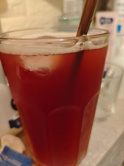 The image shows a close-up of a tall glass filled with a dark red beverage, likely iced tea or a similar drink, with ice cubes and foam on top. A dark brown stirrer or straw is visible in the drink. The setting appears to be indoors, with a blurred background that suggests a countertop or table. There is a hint of a second, empty glass to the right of the main glass. In the background, there are out-of-focus objects, including what might be a box or container with some blue and white markings. Some text is partially visible on a blue package in the lower left corner of the image, but it is too blurred to be legible. The lighting is somewhat dim, casting a warm tone over the scene. No people are visible in the frame, and the focus is entirely on the drink and its immediate surroundings.