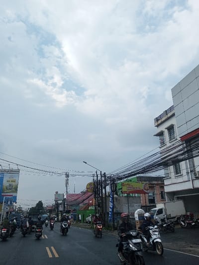 A street view under a cloudy sky shows a busy road filled with motorcycles. Buildings line the side of the road, with several billboards and power lines creating a cluttered visual landscape. A white building is visible on the right side of the image. The road has double yellow lines and the sky dominates the upper portion of the image.