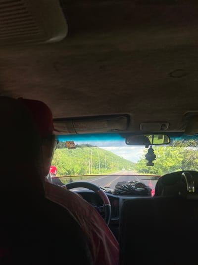 The image captures the interior of a vehicle, looking forward through the windshield at a road lined with lush green trees and rolling hills, indicative of a rural or semi-rural setting. The scene suggests a journey through San Lorenzo, Honduras, a region known for its tropical climate and scenic landscapes. The sky is partly cloudy, with a bright, sunny atmosphere.

In the foreground, a person wearing a red baseball cap and glasses is seated in the driver's seat, their back to the camera. They are wearing a pink and white striped shirt. Another individual, wearing a red helmet, is visible in the passenger seat. The dashboard of the vehicle is cluttered with various items, including a black plastic bag, a traffic cone, and a black air freshener hanging from the rearview mirror. A small yellow sticky note with writing on it is affixed to the windshield. The road ahead shows a few distant vehicles, suggesting moderate traffic. The overall impression is that of a casual road trip through a vibrant, green environment.