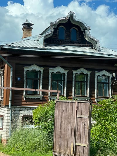 This is a vertical, eye-level photograph of a rustic wooden house in Kletino, Russia. The house features ornate, carved wooden window frames with a pale, weathered finish, and dark wooden walls. Several windows are visible, some with curtains. Above the windows, the roofline is complex with a decorative metal trim. A stone chimney with a metal cap extends upwards from the roof. To the right of the house, there is a large, weathered wooden door or gate, partially obscured by lush green foliage and tall grass. The sky above is partly cloudy with bright white clouds against a blue background. A metal address plate with the number "45" is affixed to the wall below one of the windows. The scene appears to be set during the daytime on a clear, sunny day, with the natural light casting shadows and highlighting the textures of the wood and foliage. The overall impression is one of a slightly aged but charming rural dwelling.