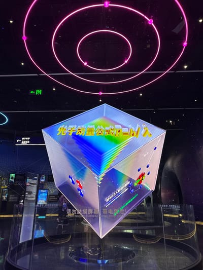 The image depicts an interior scene within a modern exhibition hall or science museum located in Guangming District, China. The focal point is a large, inverted cube-shaped digital display suspended above a dark, reflective circular base. The top surface of the cube displays the Chinese characters "光子动量公式 P=h/λ" (Photon momentum formula P=h/λ) in yellow, set against a rainbow gradient background of blue, green, yellow, orange, and red horizontal stripes. Its side surfaces show animated scientific diagrams, including representations of atoms or particles with blue rings, red spheres, and green spheres, accompanied by Chinese text such as "电子" (electron) and "波长入的入射的X射线" (incident X-ray of wavelength λ). A warning sign at the base of the cube reads "请勿触摸屏幕 带电危险!" (DO NOT TOUCH THE SCREEN. ELECTRIC SHOCK HAZARD!).

The ceiling is black, featuring concentric pink neon rings and scattered small pink spherical lights, suggesting a celestial or atomic orbital theme. A green "EXIT" sign with an arrow is visible on the ceiling to the upper left. To the left of the main exhibit, dark wall panels with blue backlighting display scientific information and diagrams. A white directional sign points left, labeled "量子科学" (Quantum Science) and "基础前沿" (Basic Frontier). Partially visible behind the main cube, a single individual wearing dark clothing stands observing the exhibits. To the right, dark, curved architectural elements or additional exhibits are dimly visible. The environment is darkly lit overall, with illumination primarily from the displays and ceiling features.