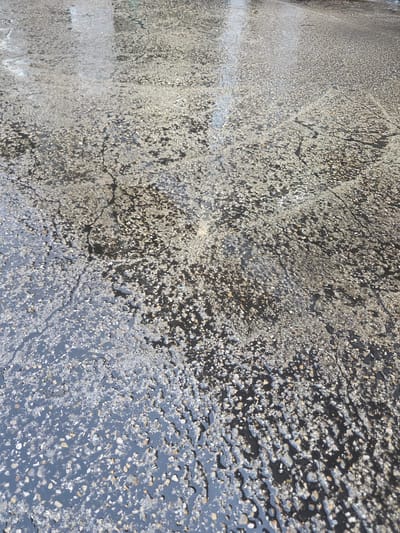 The image shows a close-up, angled view of a wet, asphalt surface. The asphalt is dark gray and speckled with small, lighter-colored aggregate stones, typical of a road or pavement. Patches of water lie on the surface, reflecting the light and creating a glossy appearance in some areas, while other areas appear drier with more visible texture from the stones. There are faint, darker lines and patterns on the asphalt, possibly indicating cracks or the way the surface has weathered. The overall impression is of an outdoor urban environment, possibly after rain. The location is described as Issy-les-Moulineaux, France.