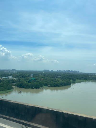 The image captures a wide, murky river flowing through a lush, green landscape in Dong Nai City, Vietnam. In the foreground, a concrete barrier marks the edge of what appears to be a bridge or elevated roadway, with a sliver of asphalt and a metal grate visible. The river itself is calm, with a few small boats, including a single, narrow vessel with a person in it, dotting its surface. The far bank is densely vegetated, with a scattering of houses and buildings interspersed among the trees. Beyond the greenery, a cityscape emerges on the horizon, characterized by a series of tall, modern buildings that contrast with the natural surroundings. The sky above is a pale blue, with scattered white clouds, suggesting a bright but possibly hazy day. The overall impression is one of a developing urban area coexisting with its natural environment.