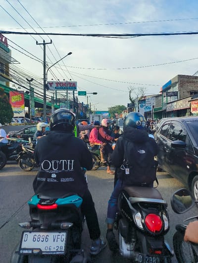 The image shows a street scene in Bojongsoang, Indonesia, located at coordinates -6.969 latitude and 107.637 longitude. The street is lined with buildings, including a shop with a "YOGYA BOJONGSOANG" sign. The sky is cloudy and the sun is shining. There are many people on motorbikes in the street, including two in the foreground wearing helmets. One of the people in the foreground is wearing a black jacket with the word "YOUTH" printed on the back. The other person is wearing a black jacket with a black backpack with a white logo. There is a car stopped in the background, and various other vehicles, including motorbikes, can be seen in the street. The street is paved and there are power lines running overhead.