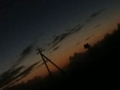 The image depicts a dark, slightly blurry twilight scene, possibly taken in Воскресенское, Russia. The sky transitions from a deep blue at the top to shades of orange and yellow near the horizon, suggesting either sunrise or sunset. Wispy clouds are visible in the sky. In the foreground, a silhouette of a utility pole with wires is prominent on the left side of the frame, angled diagonally. To the right of the pole, a dark, silhouetted tree line is visible. A small, dark rectangular signpost is faintly discernible in the middle ground, near the horizon. The overall impression is one of a rural or semi-rural landscape at dusk or dawn, with minimal man-made structures visible beyond the utility pole.