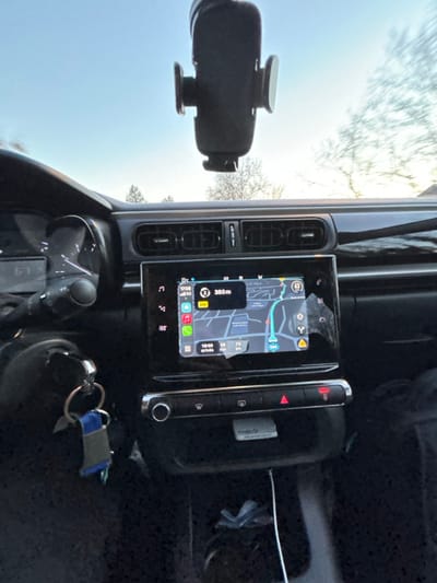 The car's interior features a prominent central display showing a map with navigation. To the left of the screen are air vents and a phone mount. A control panel with buttons is to the right of the display. Keys with a blue fob rest on the dashboard near the steering wheel and speedometer. The exterior view through the windshield shows the sky and trees.