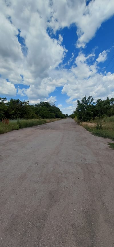 A vertical, eye-level shot captures a paved, unpaved road in Ketrysanivka, Ukraine, leading into the distance. The road is rough and cracked, with patches of gravel and dirt. On either side of the road, dense green trees and foliage line the path, with some taller trees in the background. A bright blue sky filled with fluffy white clouds dominates the upper portion of the image. In the far distance, power lines are visible against the sky. The overall scene depicts a rural, somewhat neglected road in a natural setting.