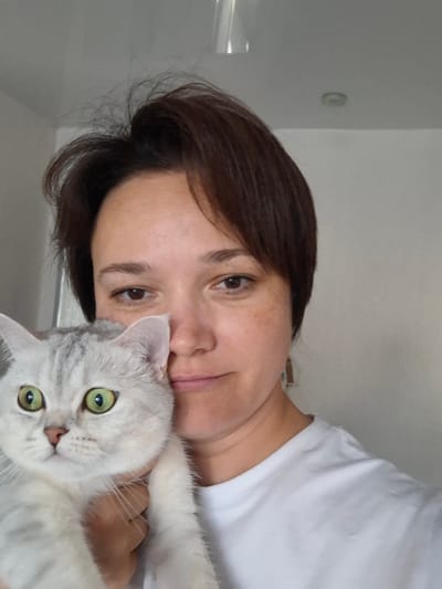 A close-up, selfie-style shot shows a woman holding a silver tabby cat in front of her chest. The woman has short, dark brown hair and is wearing a white t-shirt. She is looking towards the camera with a slight smile. The cat has bright green eyes and is looking directly at the camera. The background is a plain, light gray wall. The image was taken in Svobodny, Russia.