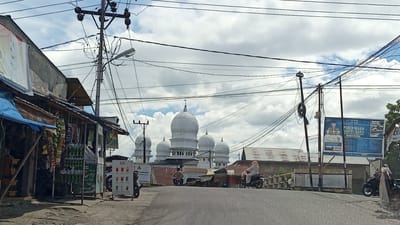 The image depicts a street scene in Aron Asan Kumbang, Indonesia, on a cloudy day. In the foreground, a paved road curves slightly to the left. On the left side of the road, a small shop with a blue tarp overhang displays various goods. Several people are visible on motorcycles. One person is riding a motorcycle on the road in the middle distance. Two other motorcycles are parked to the left of the road, and another is seen to the right.

In the background, a prominent white mosque with multiple domes dominates the skyline. Power lines crisscross the sky above the scene, supported by utility poles. A large blue billboard with Indonesian text and images is positioned to the right of the mosque, near a street light. The surrounding buildings are of varied construction, with some featuring corrugated metal roofs. The overall atmosphere suggests a typical day in a semi-urban or rural Indonesian setting.