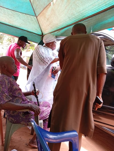 This daytime photograph, taken in Umundugba, Nigeria, depicts a group of people gathered outdoors under a light blue-green tent or canopy. The ground beneath is reddish soil.
In the foreground, an older man with dark skin is seated on a light green plastic chair to the left. He wears a purple and white patterned long-sleeved outfit and holds a black walking stick, to which a white electrical plug is visibly attached. His feet are bare. To his right, with his back to the viewer, stands a man in a long, brown traditional tunic and trousers, holding a black smartphone in his right hand.
In the center-middle ground, a woman with dark skin is standing, wearing a white headwrap adorned with pearls and a long white dress with cut-out embroidery. She also wears a pearl necklace. She appears to be interacting with a dark-colored car, partially visible on the right, which has an open door. Further back on the left, a younger man in a pink sleeveless top and black baseball cap is partially visible, also near the car.
The scene suggests an informal social gathering or a moment of arrival or departure, possibly a family event. The bright lighting indicates clear, sunny weather. No visible text is discernable in the image.
