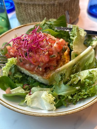 A close-up, eye-level shot shows a single serving of food on a light-colored plate with a thin, golden-brown rim. The dish consists of a slice of food, possibly a tart or pastry, topped with a diced tomato mixture and garnished with vibrant pink sprouts. The base is surrounded by a generous portion of fresh green lettuce leaves. A silver fork is partially visible on the left side of the plate, and a knife rests on the upper right. In the background, a woven basket and a blue glass bottle are out of focus, suggesting an outdoor or rustic dining setting. The lighting is natural, casting soft shadows. The scene is likely from a restaurant in Veules-les-Roses, France, given the overall ambiance and presentation.