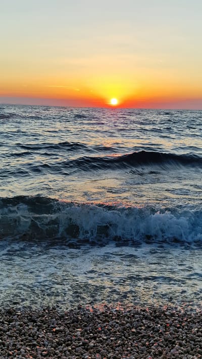 The image depicts a sunset over the Adriatic Sea in Zadar, Croatia. The sun, a vibrant orange orb, is low on the horizon, casting a warm glow across the sky and reflecting on the water. Gentle waves roll towards a pebble beach, their crests catching the golden light. The water is a deep blue, transitioning to a lighter, more agitated hue where it breaks against the shore. The beach itself is composed of dark, smooth pebbles, some of which are illuminated by the setting sun, creating small points of fiery red light. The overall atmosphere is serene and tranquil, evoking a sense of peace and natural beauty. The time of day is clearly sunset, and the weather appears to be clear and calm, ideal for enjoying a seaside view. There is no visible text in the image.