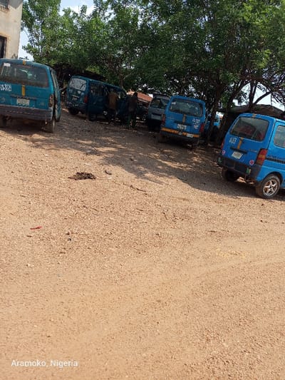 The image shows a dirt parking lot with five blue vans parked in a row. The vans are all facing the same direction, with the front of the vans facing the right of the image. Three of the vans are parked close together in the center of the image, while the other two are parked further apart at either end of the row. There are two men standing near the center of the image, beside the three vans parked together. The men appear to be talking to each other. The vans are parked in a dirt lot in Ilobu, Nigeria, at the coordinates 7.835 latitude and 4.478 longitude. The dirt lot is covered in a layer of loose sand and small pebbles. Trees line the edge of the dirt lot, casting a shadow over the parking lot. There is a building at the edge of the image, which is only partially visible. The building is painted a light gray color and has a white roof. The vans appear to be in good condition. The men are dressed in casual clothes. The sky is a bright blue. The overall scene is one of everyday life in Ilobu, Nigeria.