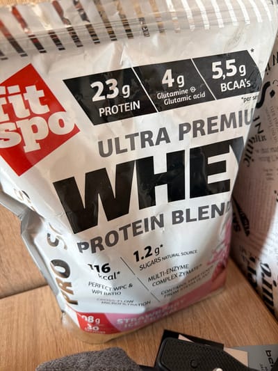 A white flexible pouch of "Fit Spo ULTRA PREMIUM WHEY PROTEIN BLEND" in Strawberry Ice Cream flavor is depicted, resting on a light brown surface, likely wood. The pouch displays prominent nutritional information, including "23g PROTEIN," "4g Glutamine & Glutamic acid," "5.5g BCAA's," "116 kcal*," and "1.2g* SUGARS NATURAL SOURCE." Text also indicates "PERFECT WPC & WPI RATIO," "CROSS-FLOW MICROFILTRATION," "MULTI-ENZYME COMPLEX ZYMATE®," and "CONTAINS SWEETENER FROM STEVIA PLANT." A weight of "798g" and "30 servings" is partially visible. To the right of the pouch, a rolled or crumpled item with black text on a white background, possibly packaging or newspaper, shows indistinct words such as "N" and "MILK." In the lower foreground, a dark grey textured object, possibly fabric, is present, alongside a black textured object and a white label with partially visible black text starting with "be...". No individuals are present, nor are there any discernible features in the image to indicate the specific location within the city of Kermen, Bulgaria, or any other geographical area.