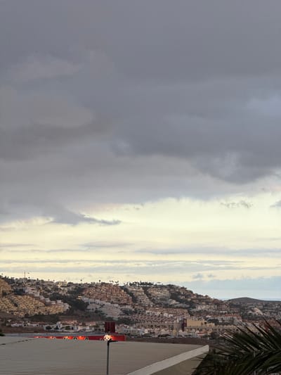This media file depicts a scenic view of a densely populated hillside town in Adeje, Spain. The town is characterized by numerous white buildings with red-tiled roofs, cascading down the slopes. In the foreground, a corrugated metal structure partially occupies the right side of the frame, with a red wall featuring a whimsical panda-shaped light fixture. A frond of a palm tree is visible at the top right edge. The sky above is dramatic, with a clear division between dark, brooding clouds on the left and bright, luminous clouds stretching towards the right horizon, suggesting a transition in weather or time of day. The presence of lights in the buildings indicates it is either dusk or dawn. The overall mood is one of natural beauty and urban development coexisting, with a hint of playful artistry in the foreground. There is no visible text in the image.