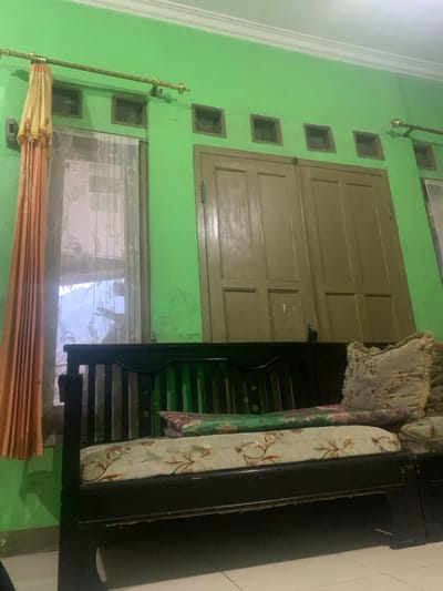 The image shows a living room in a home located at coordinates -6.928, 107.535 in the city of Lagadar, Indonesia. The room has green walls, a wooden door, a window with a sheer curtain, and a black wooden couch with floral patterned cushions. The couch is empty and there are no people in the image. The lighting is dim and the room appears to be somewhat cluttered. The window is partially obscured by the curtain, but it appears to be clear outside, as the trees and sky are visible. The door is closed, with a pattern of wood paneling. The couch is located in front of the window and the door, taking up a significant portion of the space. The walls are plain green with a white border at the ceiling.