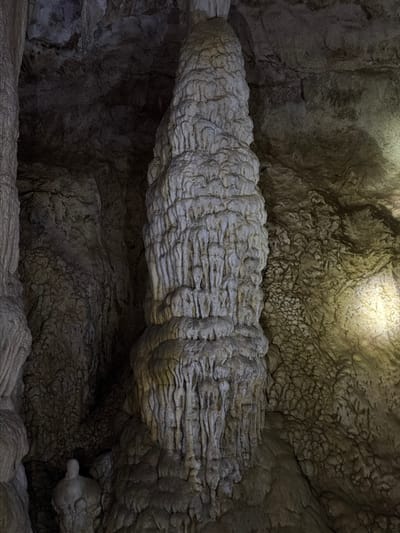 This image depicts the interior of a natural cave located in Phang-nga, Thailand. A large, light-grey geological column, consistent with a stalagmite or cave pillar, occupies the majority of the vertical frame, positioned slightly right of center. This formation displays a heavily textured, layered, and intricate surface, indicative of extensive mineral deposition. The surrounding cave walls and ceiling are dark, rough, and uneven, featuring various smaller, irregular rock formations and additional vertical structures on the left periphery. A smaller, rounded geological formation is discernible at the base of the main column on the left. A distinct yellowish artificial light source illuminates the lower-right section of the primary column and an adjacent textured wall, creating localized highlights against a predominantly shadowed background. No human figures or man-made objects are visible. The scene is static, documenting natural subterranean geological features.