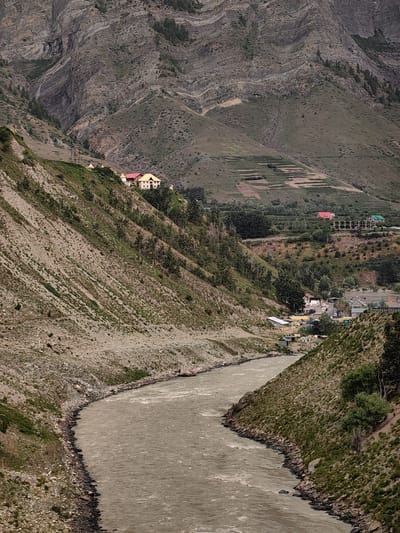 The image depicts a wide, muddy river flowing through a valley in Gondhla, India. Steep, rocky mountains with sparse vegetation rise on either side of the river. On the left bank, the mountainside is characterized by loose rocks and patches of dry grass. On the right bank, the terrain is more vegetated, with trees and shrubs dotting the slopes.
In the middle ground, a small settlement is visible nestled in the valley. Several buildings with colorful roofs, including a prominent building with a yellow roof and red trim, are situated on the hillside. Further into the valley, more structures, possibly houses and shops, are clustered together, with a road winding through them. Some tents are also visible near the settlement.
In the distance, terraced fields are carved into the mountain slopes, indicating agricultural activity. The sky is overcast, casting a diffused light over the scene. The overall impression is one of a remote, mountainous region with a small human presence integrated into the natural landscape.