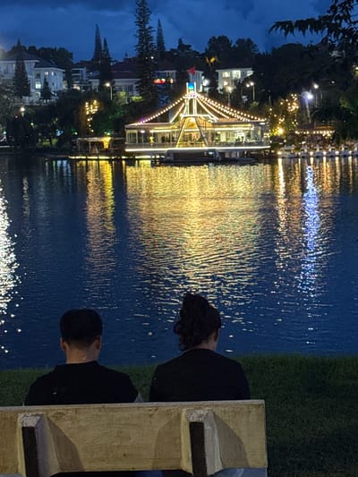 In Xuan Huong Lake, Da Lat, Vietnam, two individuals are seated on a concrete bench facing the water. The scene depicts a nighttime setting with the lake reflecting the illuminated buildings and decorative lights from a structure across the water. The structure, possibly a restaurant or entertainment venue, is adorned with strings of golden lights, and further lights are visible on the adjacent buildings and trees on the hillside. The water's surface is disturbed by ripples, causing the reflections to appear elongated and shimmering. The foreground includes a grassy area and the bench on which the two individuals are seated. Their backs are to the camera, so their features are not visible.