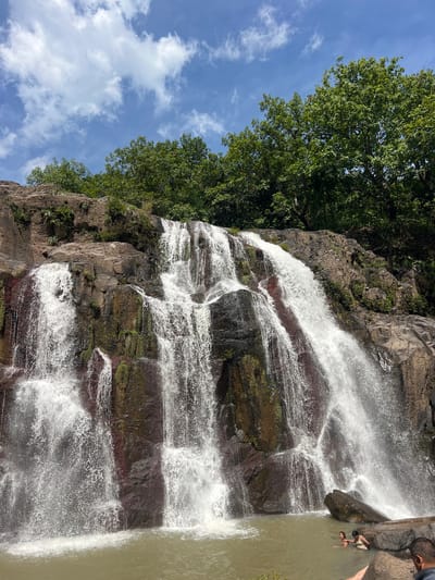 A wide shot shows a multi-tiered waterfall cascading down a rocky cliff face. The water is white and frothy as it falls, creating mist at the bottom. Lush green trees top the cliff, and the sky above is a bright blue with scattered white clouds. In the foreground, a body of murky water collects at the base of the waterfall. Two women are partially submerged in the water, and the head and shoulders of a man are visible on the right side of the frame, in the lower foreground. The scene appears to be the El Salto waterfall in Arambala, El Salvador.