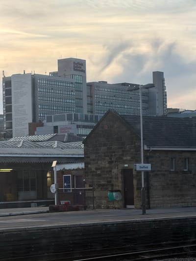 The image shows a view of Sheffield Hallam University and Sheffield Train Station. It's a cityscape with modern university buildings in the background, and a brick-built train station platform in the foreground, including tracks. A sign indicates the station is Sheffield Station. The sky appears overcast.