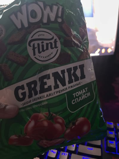 The scene, set indoors in Karmana, Uzbekistan, captures a close-up, angled view of a green and white "Flint" brand snack package. The package prominently displays "WOW!" at the top, followed by the brand "Flint" with "2002" and "СУХАРИКИ" (croutons) in a white oval logo. The product name "GRENKI" (croutons) is in large lettering, with "JAVDAR GRENKILARI/ГРЕНКИ РЖАНЫЕ" (Rye Grenki) below it. A green rectangular label on the right indicates the flavor "ТОМАТ СПАЙСИ" (Tomato Spicy). Illustrations of dark brown croutons and three red tomatoes with some reddish flakes are visible on the package. A human thumb and a portion of an index finger, displaying light to medium skin tone, are positioned on the lower-left, holding the snack bag. Beneath the package and to the right, a black mechanical keyboard with blue backlighting on its keys is partially visible. The background is blurred, showing indistinct bright light sources, suggesting a screen or window in an otherwise dimly lit interior.